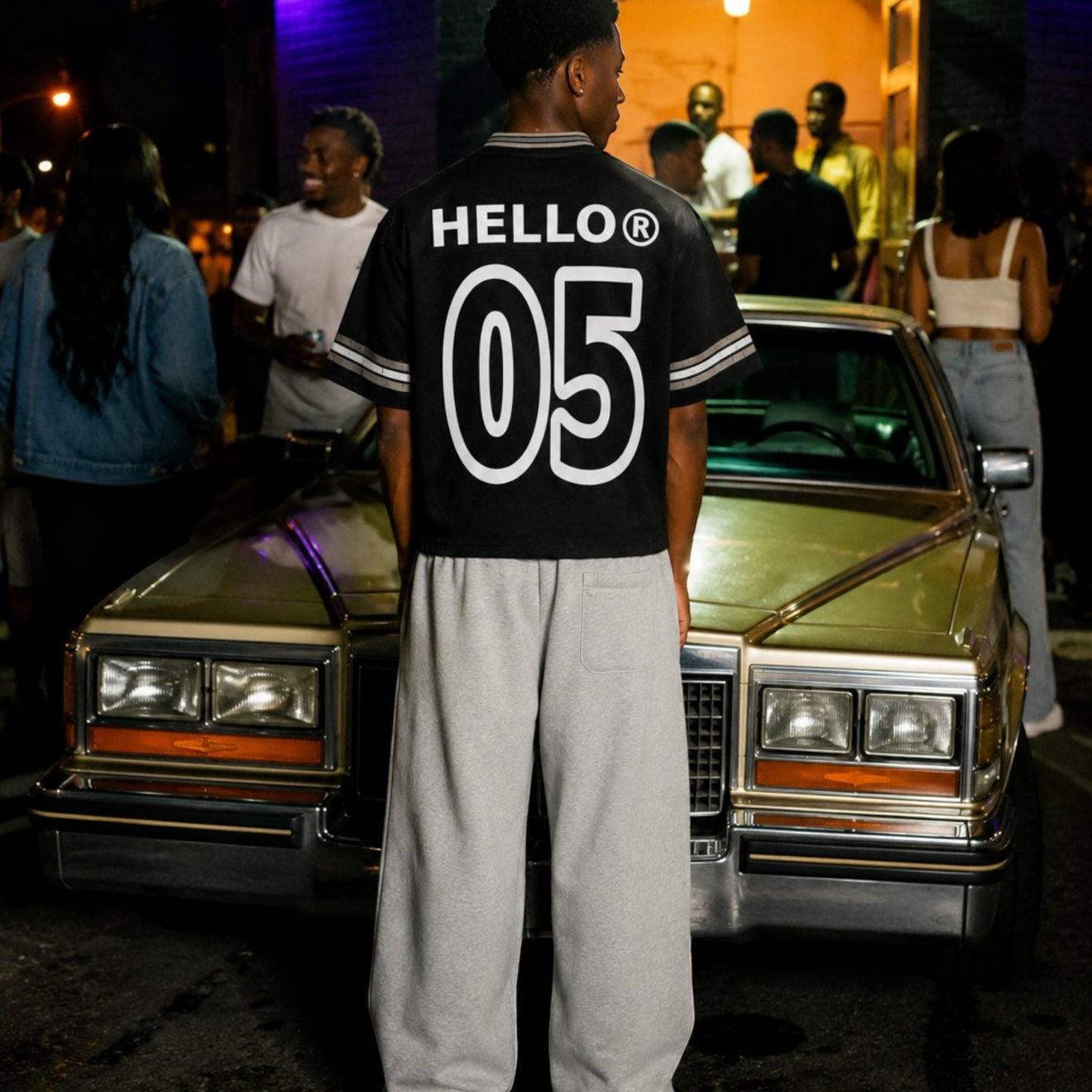 Footprint Apparel men's streetwear — man with his back to the camera in a black HE★LO 05 jersey in front of a vintage gold Cadillac at night