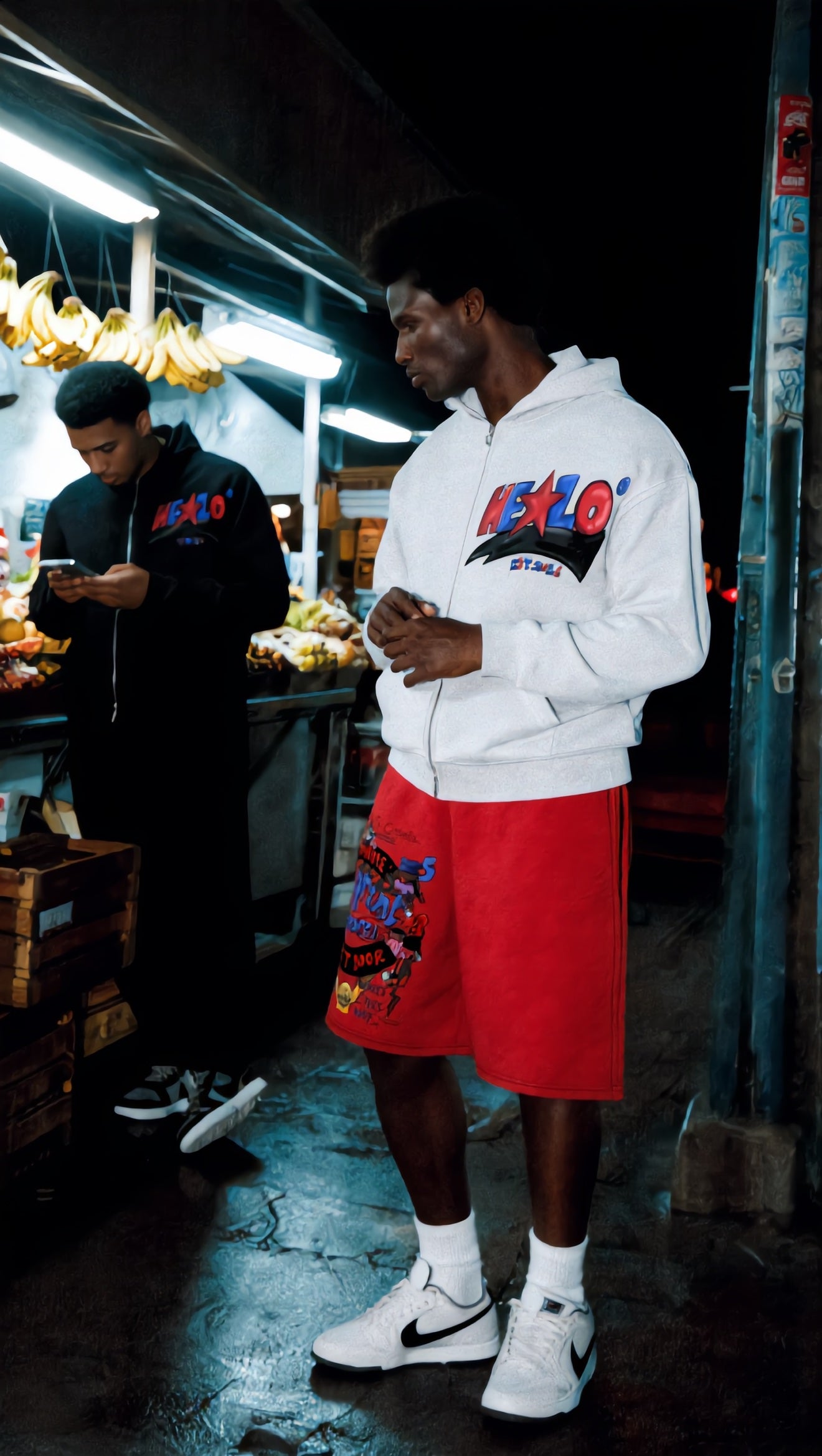 Footprint Apparel — man in the white HELLO zip hoodie and red shorts at a night market bodega, wide shot