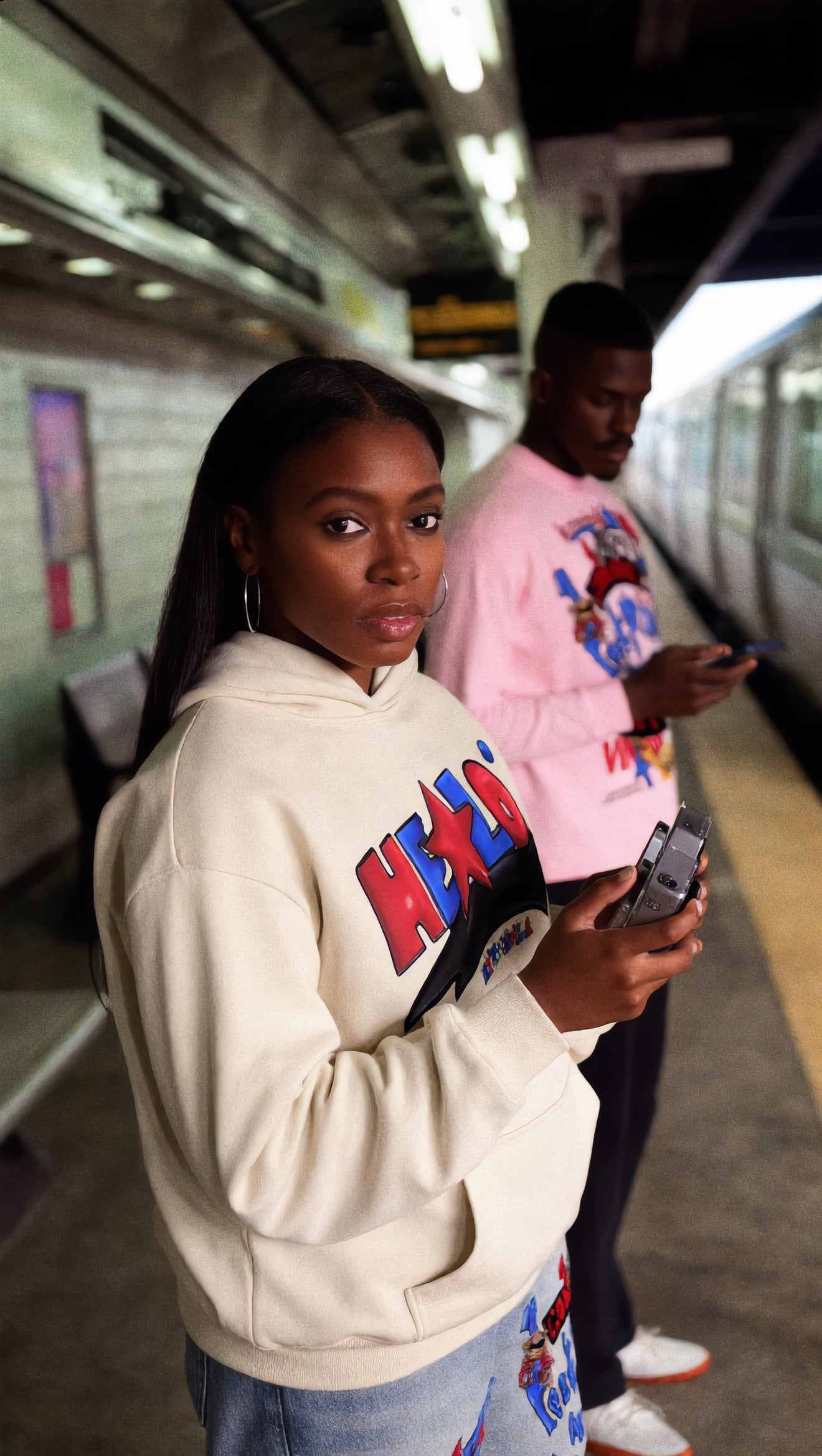 Footprint Apparel — woman in front wearing the cream HE★LO hoodie on a subway platform, man in pink behind
